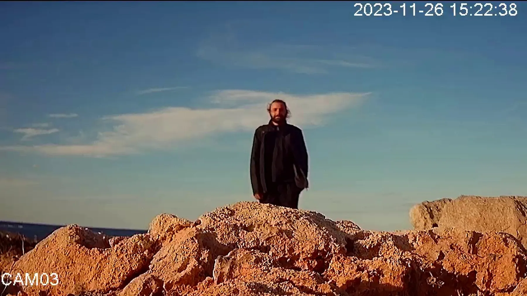 CCTV image of a person standing behind rocks beneath a blue sky.