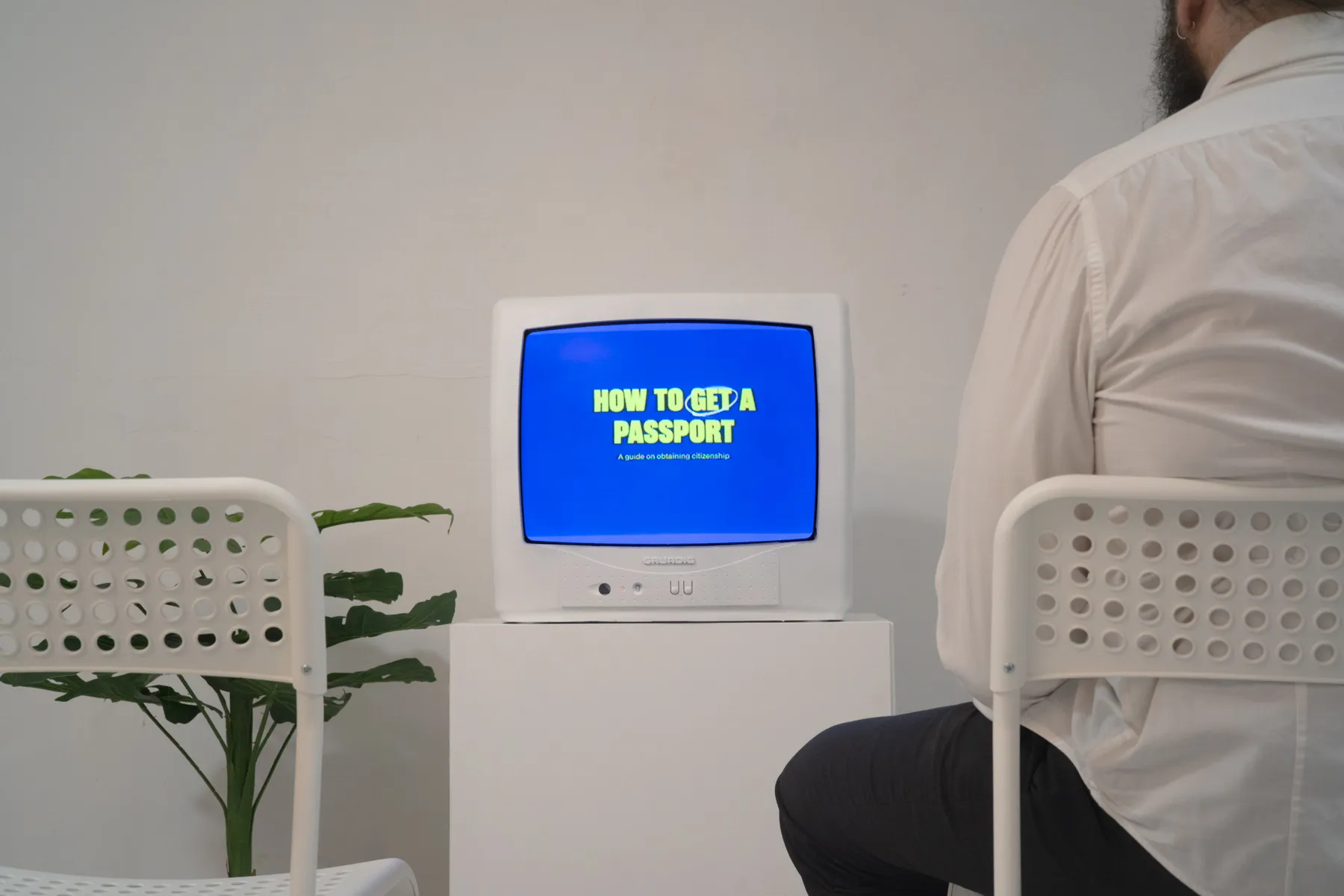 Viewer seated in the installation waiting room, facing a television displaying How to get a passport.