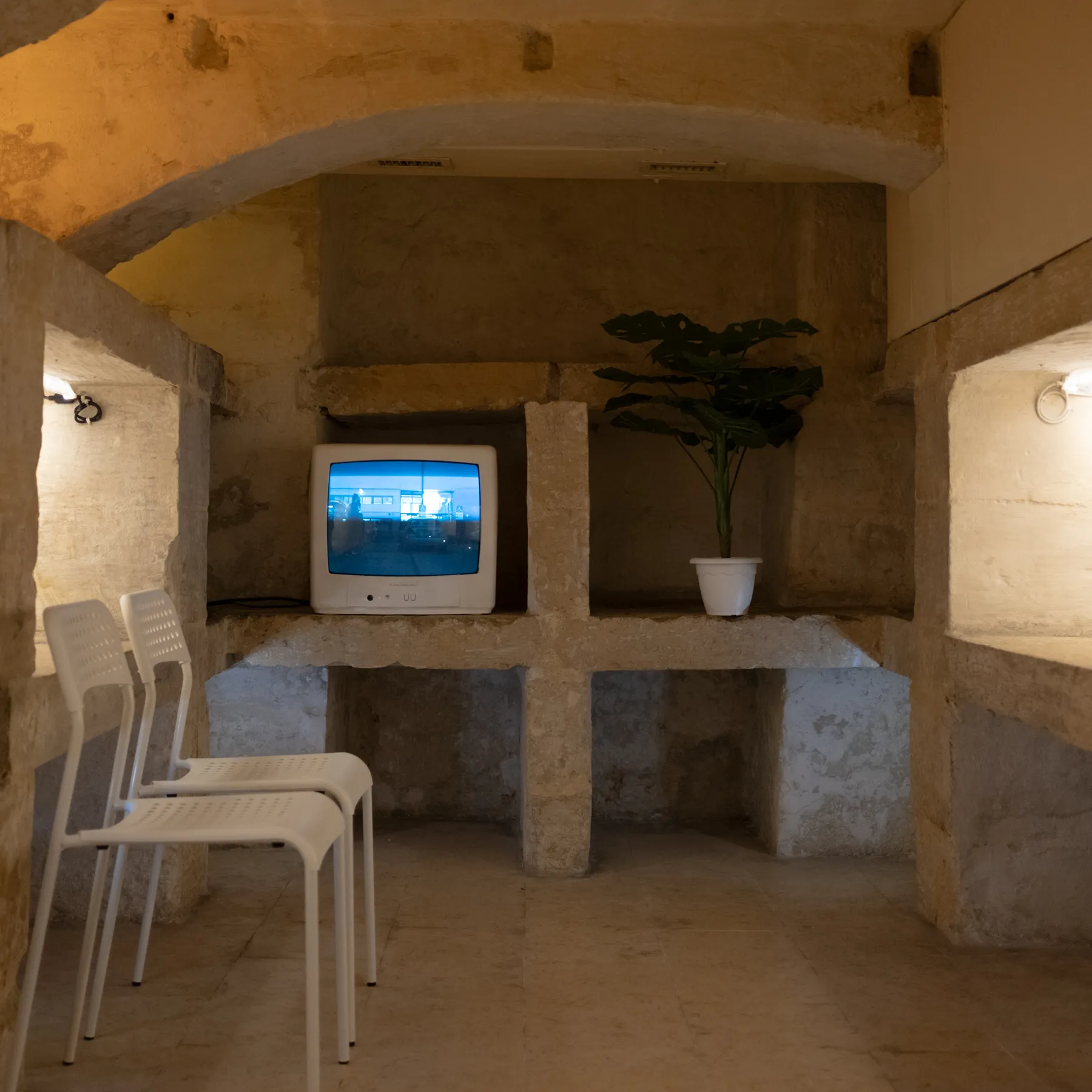 Installation view with waiting room chairs, a television and a potted plant in a stone interior.